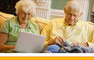 Photo of two seniors; the woman is using a laptop and the man is knitting